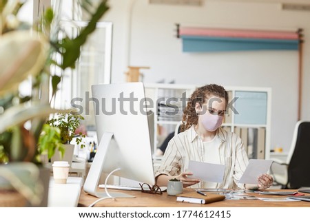 Front view portrait of young female photographer wearing mask while reviewing pictures sitting at desk in home studio, copy space