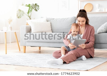 Smiling Mother And Her Adorable Baby Son Watching Cartoons On Digital Tablet At Home Together, Sitting On Floor In Living Room