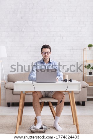 Work from home and humor. Busy, serious guy in shirt and shorts works at laptop and puts foot on dumbbell in interior of living room, free space