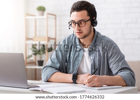 Teacher online. Guy in glasses and headset makes notes in notebook and looks at laptop in living room interior, copy space