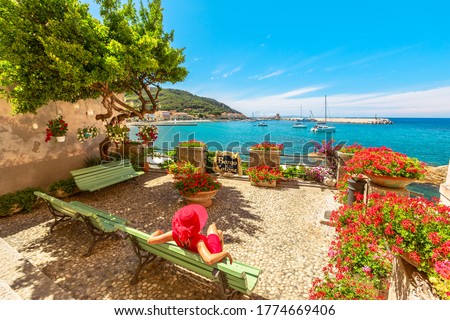 Lifestyle woman in red sitting on bench in Marciana Marina, flowery old district Borgo al Cotone: meaning COTTON VILLAGE, looking harbor bay with ships. Tourist on holiday travel in Italy, Elba island Royalty-Free Stock Photo #1774669406