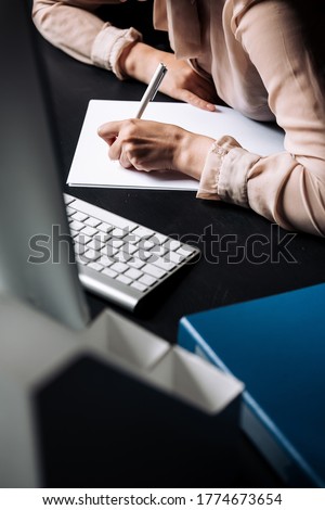 Close-up of a girls left-handed girls hand who writes on white sheets while sitting at a table in an office or library.
