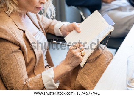 Contemporary mature businesswoman with pen and notebook making working notes or planning work while sitting by desk