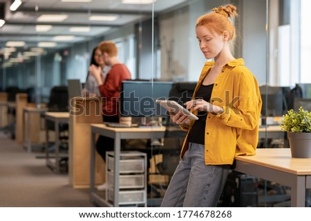 Pretty young businesswoman in casualwear making presentation in digital tablet while leaning on desk with green plant in flowerpot