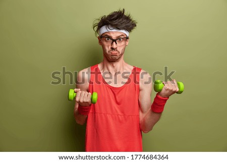 Motivated determined sportsman leads sporty lifestyle, lifts heavy dumbbells for muscle training, does morning fitness at home, wants to have biceps, wears red shirt and wristband, looks sadly