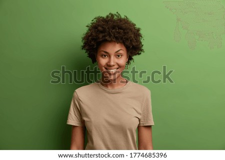 Headshot of cute smiling ethnic woman with Afro hair grins at camera and enjoys good summer day, looks unbothered and happy, gives sincere friendly smile to you, stands against green background
