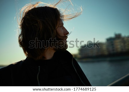 Mysterious romantic young Caucasian bearded man with hair flying in wind posing outdoors, walking on embankment on sunny day, looking on lake and cityscape in background. Picture against sun