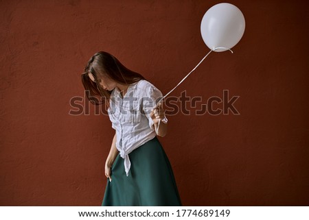 Isolated studio image of beautiful young woman with long loose brown hair posing against blank wall background, holding white helium balloon, celebrating birthday, looking down, hiding face