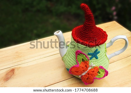 Decoration for the teapot made of multi-colored yarn in the form of a butterfly knitted manually on a white teapot, against the background of a cup of tea. Everything stands on a natural wooden table.