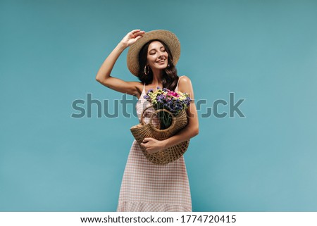 Cheerful modern brunette with earrings and cool hat in stylish light dress posing with straw handbag and colorful flowers..