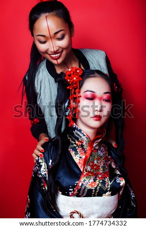 two pretty geisha girls friends: modern asian woman and traditional wearing kimono posing cheerful on red background
