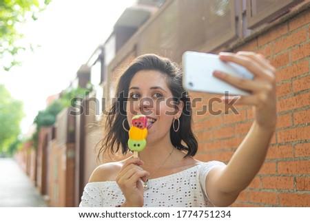 Photo of a young and attractive woman eating an ice cream and taking a selfie with her phone with an orange background
