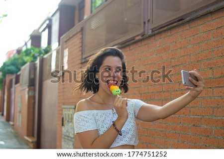 Photo of a young and attractive woman eating an ice cream and taking a selfie with her phone with an orange background