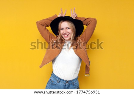 young girl in autumn jacket and hat shows a peace sign and smiles on a yellow isolated background