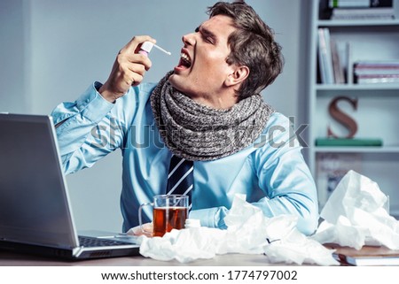 Sick employee using spray for throat. Photo of young man in office suffering virus of flu. Medical concept. Royalty-Free Stock Photo #1774797002
