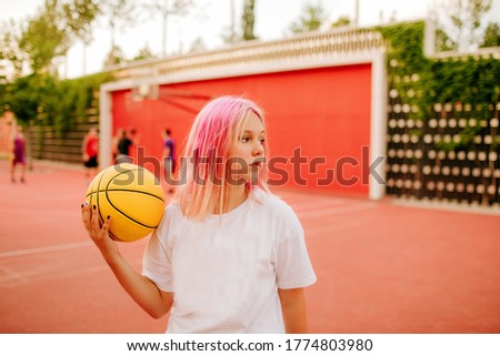A beautiful teenage girl with dyed pink hair and a white t shirt stands on a basketball court against a background of basketball players and holds a yellow basketball ball