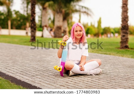 A beautiful teenage girl with pink hair in a white t shirt and white socks sits on the ground and holds a colored skate