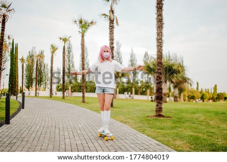 A beautiful teenage girl with pink hair in a white t shirt , white socks and medical mask rides a skateboard in the background of the stadium