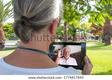 Rear view of a senior woman sitting in public park in online chat with a smiling attractive friend or husband - talking, chatting, communicating, two retirees stay in touch remotely.
