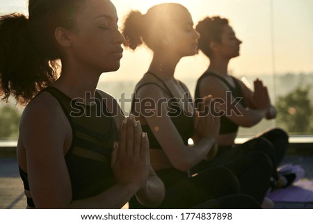 Calm serene sporty young african american ethnic woman meditate sit namaste eyes closed doing breathing exercises practice relaxing at outdoor group multiethnic yoga class in morning sunrise light.