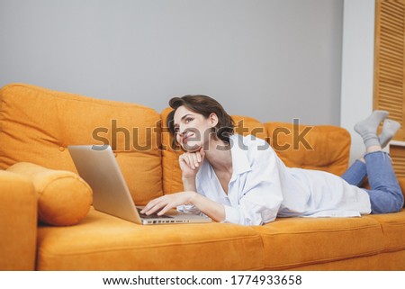 Pensive young brunette woman in casual clothes lying on couch spending time in living room at home. Rest relax good mood leisure lifestyle concept. Mock up copy space. Working on laptop pc computer