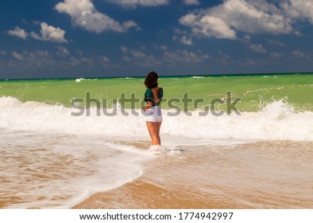 girl is standing on wavy sandy beach with her green swimsuit and white short. Blue sky with natural white clouds 