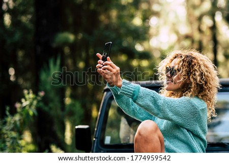 Outdoor people enjoy technology with cheerful caucasian woman take a picture with smartphone in the forest during a travel with her car - adult female and alternative vacation with nature