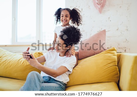 Black young cheerful woman taking selfie with laughing funny kid with pigtails behind back on yellow home couch with pillows