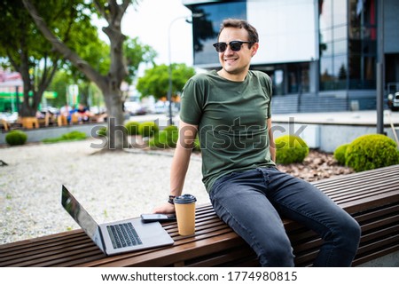 Handsome young man spending time outdoors at the city, using laptop computer while sitting on a bench and drinking takeaway coffee