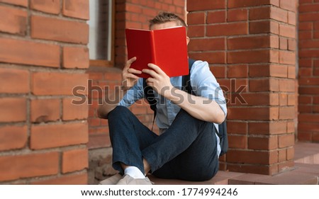 European male student sitting near the library and reading a book. Self-education concept