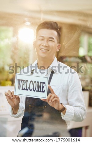 Portrait of happy excited young coffeeshop owner sticking sign on entrance door and welcoming customers inside