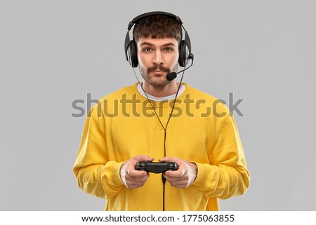 technology, gaming and people concept - young man or gamer in headphones with gamepad playing and streaming video game over grey background