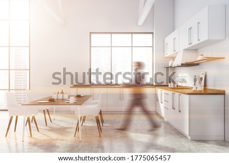 Blurry young man walking in modern kitchen with white brick walls, concrete floor, white countertops and dining table with chairs. Toned image
