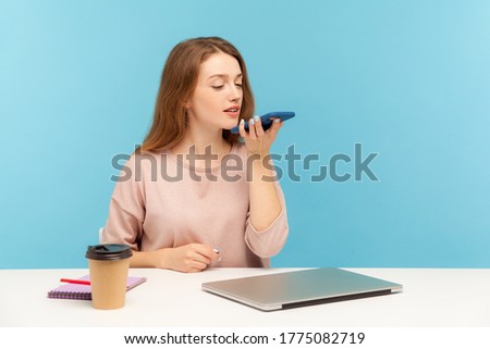 Digital speaker app. Young woman employee sitting at workplace and talking to mobile phone, using voice assistant to record reminder of business event. indoor studio shot isolated on blue background