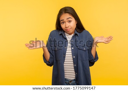 Have no idea, maybe! Portrait of confused girl in denim shirt shrugging shoulders in bewilderment, doubting and feeling uncertain, don't know answer. indoor studio shot isolated on yellow background Royalty-Free Stock Photo #1775099147