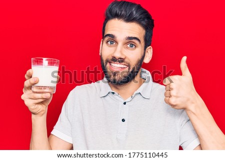 Young handsome man with beard drinking glass of milk smiling happy and positive, thumb up doing excellent and approval sign 