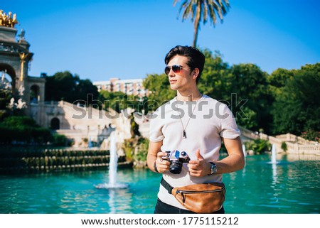 Handsome caucasian male photographer amateur spending vacation in city standing on historical place with fontagne, trendy dressed man tourist with camera exploring town destinations on sunny day
