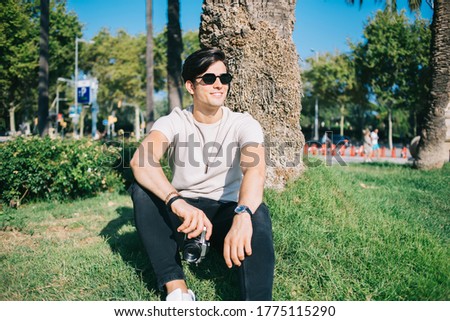 Cheerful caucasian male photographer in sunglasses enjoying sunny day leisure in park sitting on grass, happy 20s hipster guy traveler holding camera for taking photos of city visiting locations