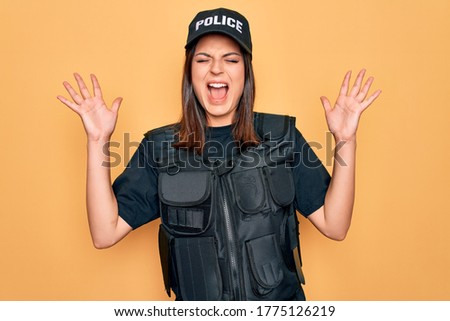 Young beautiful brunette policewoman wearing police uniform bulletproof and cap celebrating mad and crazy for success with arms raised and closed eyes screaming excited. Winner concept