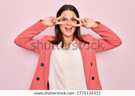 Young beautiful brunette businesswoman wearing elegant jacket over isolated pink background Doing peace symbol with fingers over face, smiling cheerful showing victory