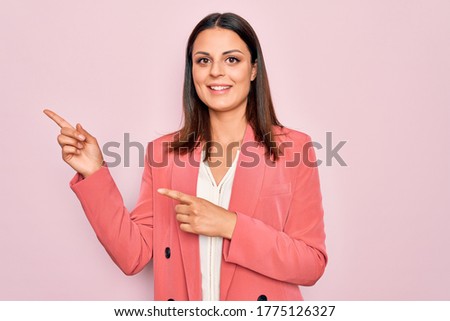 Young beautiful brunette businesswoman wearing elegant jacket over isolated pink background smiling and looking at the camera pointing with two hands and fingers to the side.