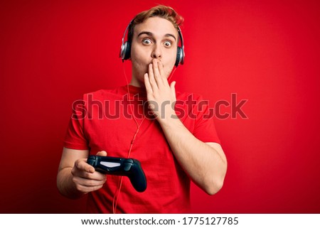 Young handsome redhead gamer man playing video game using headphones and joystick covering mouth with hand, shocked and afraid for mistake. Surprised expression