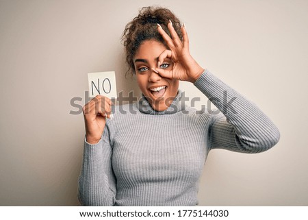 Young african american girl holding reminder paper with no word negative message with happy face smiling doing ok sign with hand on eye looking through fingers