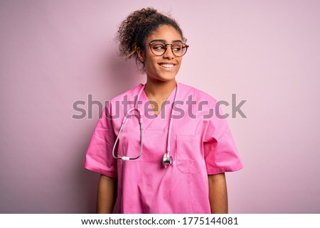 African american nurse girl wearing medical uniform and stethoscope over pink background looking away to side with smile on face, natural expression. Laughing confident.