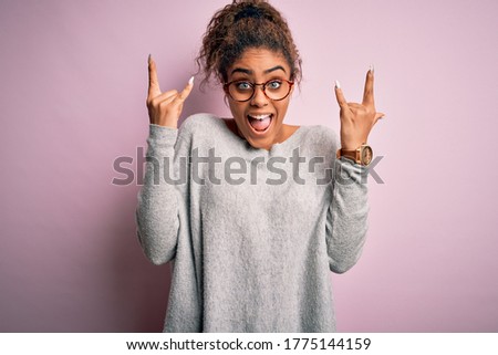 Young beautiful african american girl wearing sweater and glasses over pink background shouting with crazy expression doing rock symbol with hands up. Music star. Heavy music concept.