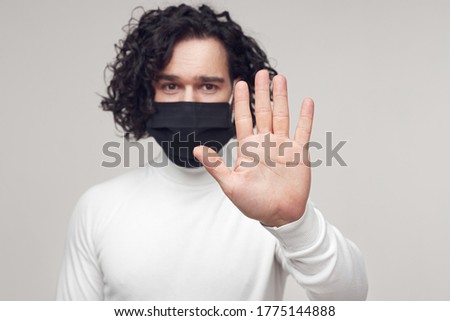 Young guy in black medical mask looking at camera and showing stop gesture while trying to prevent coronavirus spreading against gray background
