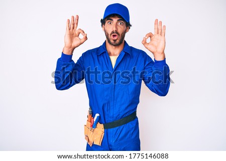Handsome young man with curly hair and bear weaing handyman uniform looking surprised and shocked doing ok approval symbol with fingers. crazy expression 