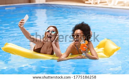 Two happy millennial girls taking selfie together in pool on summer vacation