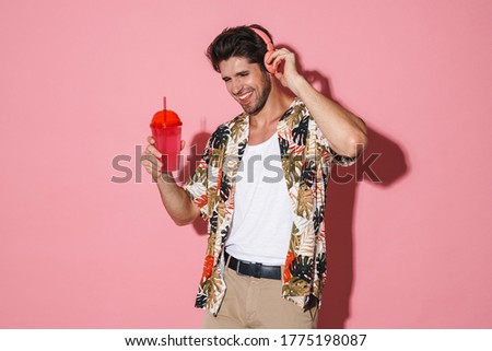 Portrait of cheerful young man using wireless headphones while drinking soda isolated over pink background