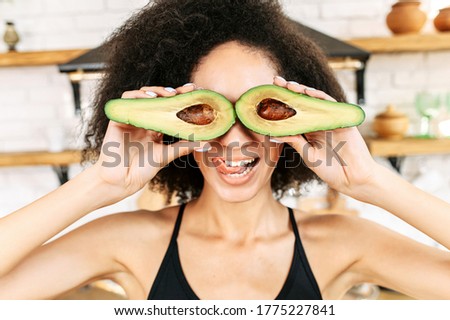 Attractive young multi ethnic woman with an afro hairstyle covered her eyes with halves of avocado. Funny picture with healthy food. Keto diet concept
